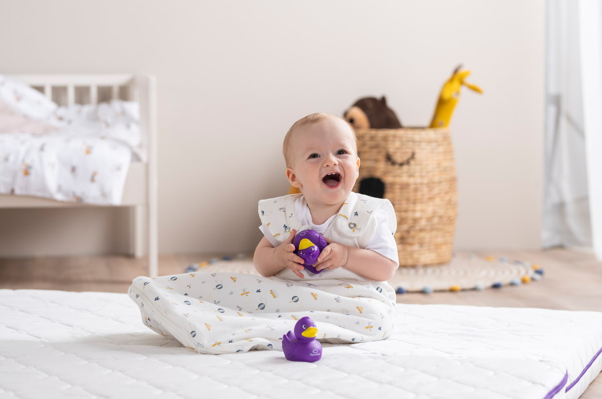 Baby im Träumeland Schlafsack sitzend auf Matratze mit Quietsche-Ente Baby im Träumeland Schlafsack sitzend auf Matratze mit Quietsche-Ente